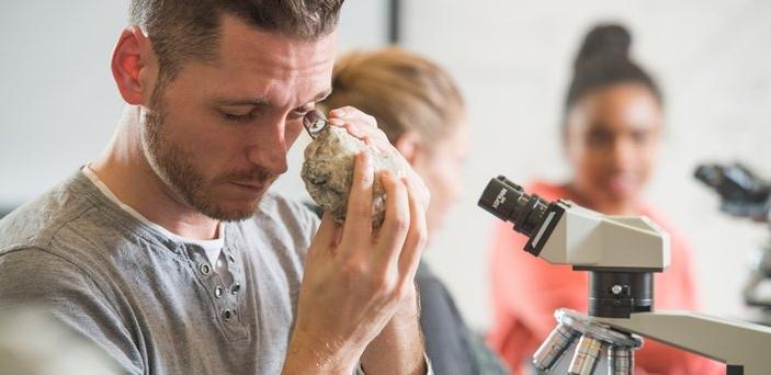 A student sitting in front of a microscope looking at a rock with a magnifier