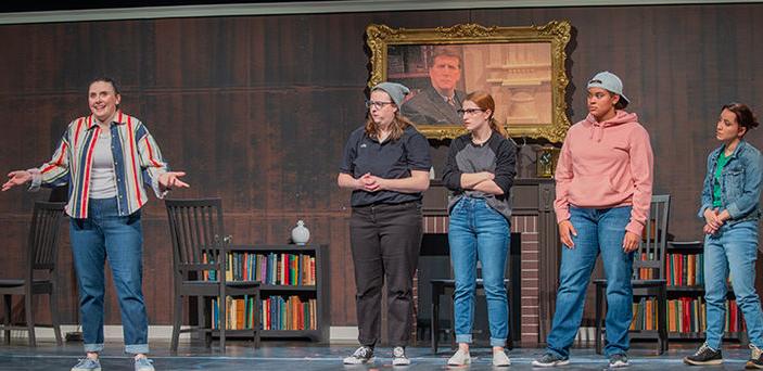 5 actors on a stage with a backdrop of a fireplace and bookcases. One actor is separate from the others, speaking and gesturing while the others look on.