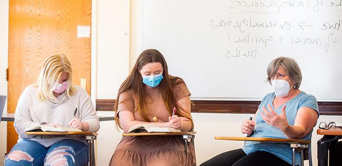 Dr. 安·布伦杰斯(Ann Brunjes)坐在讲台上讲课，面前是一块白板，上面显示着部分笔记，上面写着“对应”和“道德和精神真理的外化”.两个学生坐在她旁边的桌子上做笔记.