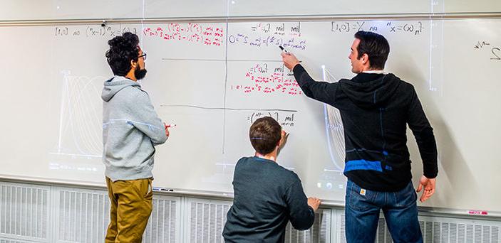 马特·萨洛蒙教授指着一个学生写在白板上的方程的一部分，这个学生手里拿着记号笔站在那里听教授讲课，而另一个学生在黑板上写方程