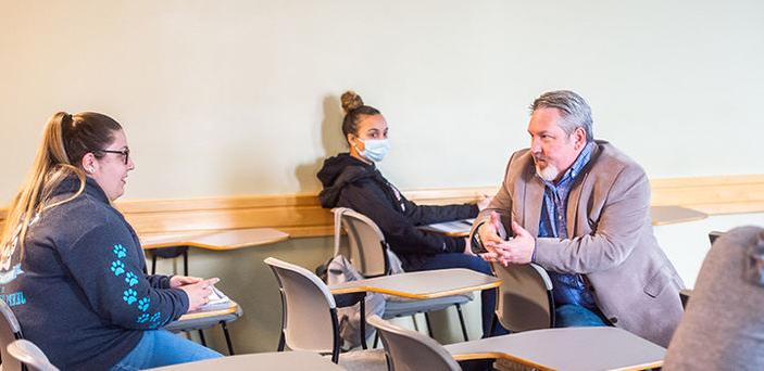 Professor Michael Devalve sitting in a classroom desk talking with a student in class
