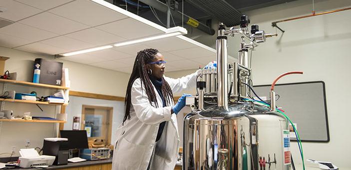 a female BSU student wearing lab coat, rubber gloves and safety glasses stands on a step stool to reach the top of a large silver piece of equipment