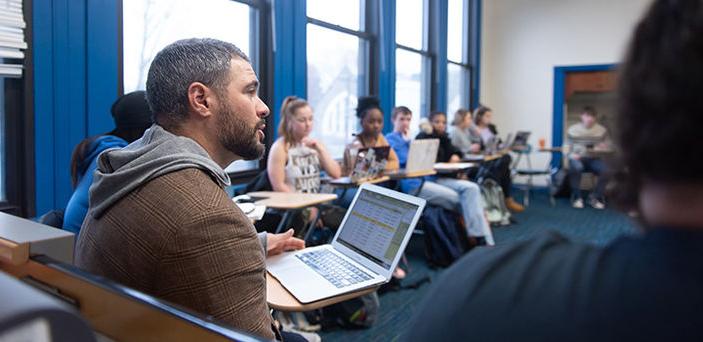 Justin Caouette教授开着笔记本电脑讲课，他坐在一张桌子上，周围是一圈开着笔记本电脑的学生. 